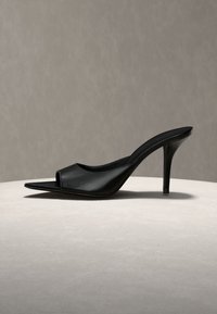Μαύρο mule με ψηλό τακούνι, μυτερή μύτη και ανοιχτή φτέρνα, εκτεθειμένο σε φωτεινή στρογγυλή επιφάνεια με ουδέτερο φόντο.