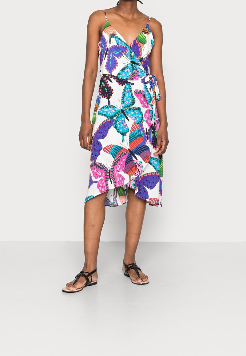 Femme portant une robe portefeuille sans manches avec des motifs colorés de papillons et de feuilles, associée à des sandales plates noires, debout devant un fond blanc.
