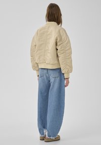 Beige gesteppte Jacke mit gepufften Ärmeln, gerippten Bündchen und tailliertem Saum, kombiniert mit locker sitzenden, zerstörten blauen Jeans und braunem Schuhwerk.
