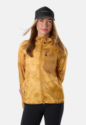 Kvinde iført en sennepsgul lynlåstrøje med hætte og sort kasket, stående med hænderne bag ryggen mod en lys baggrund.