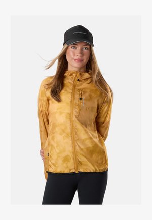 Kvinde iført en sennepsgul lynlåstrøje med hætte og sort kasket, stående med hænderne bag ryggen mod en lys baggrund.