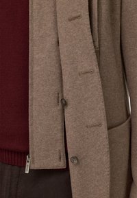 Brązowy kardigan z mieszanki wełny o fakturowanym wykończeniu, z detalami guzików i przednią kieszenią, noszony na ciemnoczerwonym swetrze.