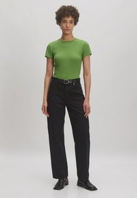 Groen aansluitend t-shirt met korte mouwen, gecombineerd met hoge zwarte broeken en zwarte schoenen, staand op een neutrale achtergrond.