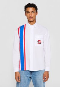Hvid langærmet skjorte med blå og røde striber på den ene side, en brystlomme med et sort og rødt grafisk patch samt knaplukning.