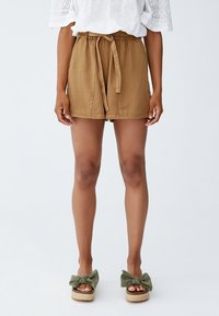 Shorts marron à taille nouée, poches latérales et coupe décontractée, associés à des sandales à enfiler vertes avec un détail nœud. Haut blanc à manches en dentelle visible.