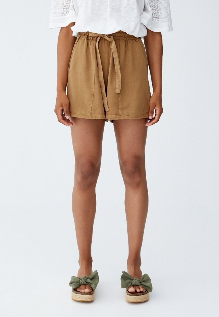 Shorts marron à taille nouée, poches latérales et coupe décontractée, associés à des sandales à enfiler vertes avec un détail nœud. Haut blanc à manches en dentelle visible.
