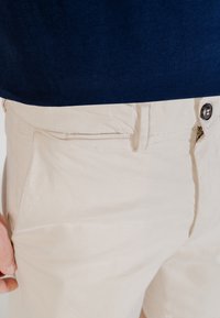 Pantalons en coton beige avec une texture lisse, dotés de poches obliques et d'une fermeture à bouton. Associés à une chemise bleu foncé.