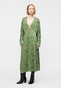 Μακρύ, πράσινο σατέν φόρεμα με δέσιμο μπροστά, διακοσμημένο με λεπτά λουλουδάτα σχέδια και μακριά μανίκια. Φοριέται με μαύρα μπότες με τακούνια.