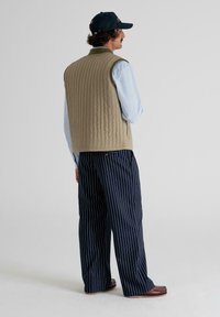 Man wearing a beige quilted vest, light blue shirt, navy pinstripe wide pants, brown shoes, and a black cap, standing facing sideways.