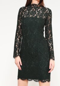 Robe en dentelle verte à manches longues, avec un col haut, un motif floral, un ourlet en scallop et des accents en dentelle transparente contrastants.