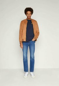 Brun suede bomberjakke, navy t-shirt med logo, blå jeans og hvide sneakers. Model står lige med hænderne i lommerne.