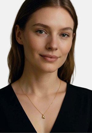 Junge Frau mit heller Haut und braunen Haaren, die ein schwarzes Oberteil und eine goldene Halskette mit einem kleinen abstrakten Anhänger trägt.