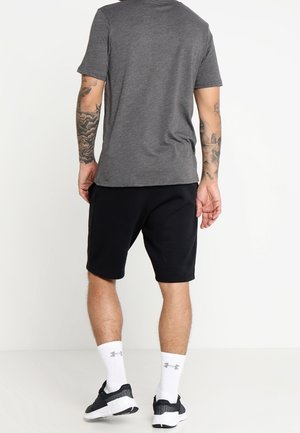Homme aux bras tatoués portant un t-shirt gris, un short noir, des chaussettes blanches Under Armour et des chaussures de sport noires, debout de dos.