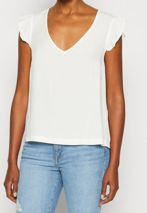 Femme portant un top blanc sans manches à col en V avec des épaules volantées et un jean bleu clair déchiré, visible du cou aux hanches.