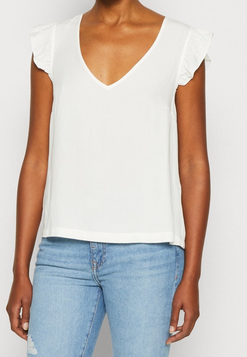 Femme portant un top blanc sans manches à col en V avec des épaules volantées et un jean bleu clair déchiré, visible du cou aux hanches.