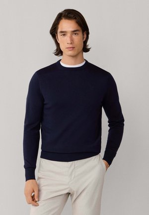 Suéter azul marino de cuello redondo con puños y dobladillo acanalados. Textura suave y lisa. Llevado sobre una camisa blanca, combinado con pantalones de color claro.