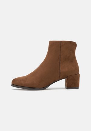 Bottine en daim marron avec un petit talon bloc, un bout arrondi et des surpiqûres latérales sur fond gris.