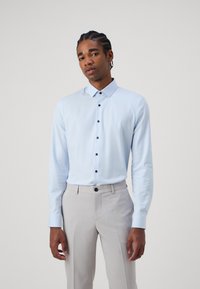 Cămașă lungă cu mâneci în culoare albastru deschis, cu guler tip button-down și nasturi închisi la culoare, asortată cu pantaloni gri deschis. Material neted, croială ajustată.