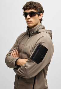 Puffjacka i taupe, med huva, dragkedja framtill och en svart ficka på vänster ärm med logotyp. Modellen bär mörka solglasögon.