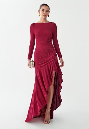 Žena v dlhých rukávoch, červenej šaty po zem s volánovým vysokým rozparkom, drží malú kovovú kabelku a má zlaté náušnice a lodičky.