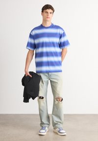 Camiseta de rayas azules con mangas cortas, combinada con unos vaqueros claros desgastados y zapatillas azules. Sosteniendo un sudadera negra.