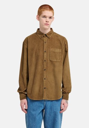 Young person with short red hair wearing a brown button-up corduroy shirt and blue jeans standing against a plain background.