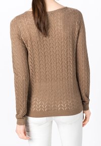 Frau mit braunem Pferdeschwanz, die einen braunen Strickpullover mit gewelltem Spitzenmuster und weißen Hosen trägt, von hinten gezeigt.