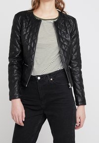 Veste en cuir matelassé noire avec poches zippées et col côtelé, portée sur une chemise rayée, accompagnée de jeans noirs taille haute.