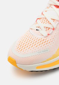 Zapatilla de running Nike con una parte superior de malla en rosa claro, acentos naranjas, cordones blancos y puntera reforzada, que cuenta con una suela texturizada para mayor tracción.