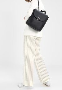 Černý kožený batoh s klopovým uzavřením, jemným detailem loga, dvěma nastavovacími popruhy a čtvercovým tvarem.