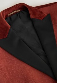 Ein roter Samtblazer mit schwarzen Satinrevers. Er verfügt über ein strukturiertes Muster am Kragen und ein glattes Innenfutter, das einen kontrastierenden Abschluss schafft.