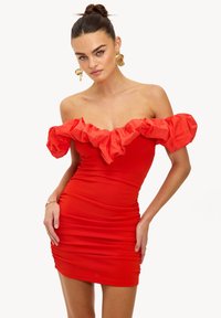 Robe rouge à épaules dénudées avec un col à volants, un corps ajusté et une texture froncée. Des boucles d'oreilles dorées imposantes complètent le look.