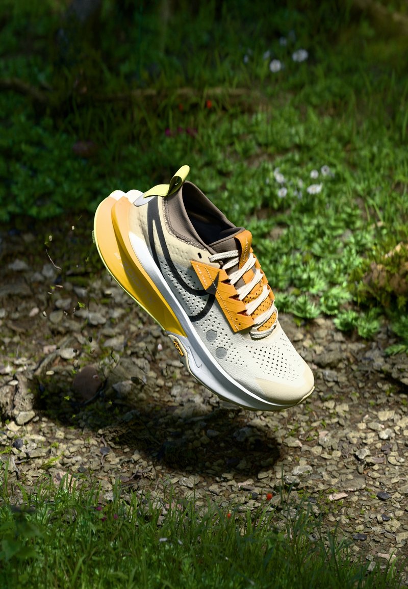Chaussure de course beige et jaune avec le swoosh noir, lacets noués, flottant au-dessus de gravier et d’herbe dans un environnement extérieur.