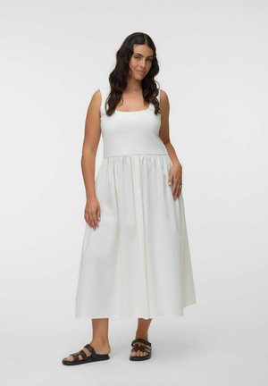Femme aux longs cheveux foncés portant une robe midi blanche sans manches et des sandales plates noires, debout devant un fond blanc uni.