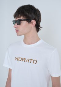 Camiseta de algodón blanca con la palabra "MORATO" en un diseño con textura de corcho marrón. El modelo lleva gafas de sol rectangulares negras.