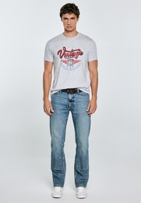 Pilkas marškinėliai su "Vintage Jeans 79" grafika, dėvimi su šviesiai mėlynomis džinsinėmis kelnėmis ir baltais sportbačiais. Atsitiktinis siluetas ir laisvas dizainas.