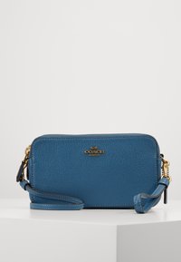 Blue leather crossbody bag with textured finish, gold logo, zippered closure, and detachable strap. Compact rectangular shape.