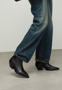Zwarte leren enkellaarzen met een puntige neus en een chunky hak, gedragen met wijd uitlopende blauwe spijkerbroeken. Soepele textuur en minimalistisch ontwerp.