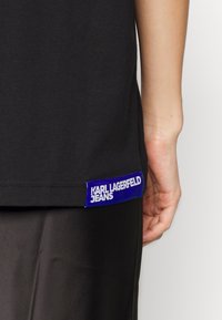 Camiseta negra con una etiqueta azul en el dobladillo que muestra "KARL LAGERFELD JEANS". Cuello redondo estándar y mangas cortas. Tela de algodón.