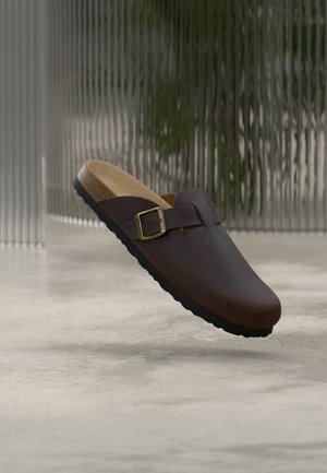 Bruine leren instapklomp met verstelbare gesp en kurken voetbed, zwevend boven een gladde betonnen vloer met een achtergrond van gestructureerd glas.