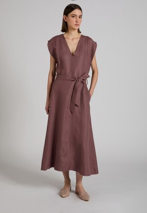 Kobieta ubrana w bezrękawową sukienkę midi w kolorze mallowym z dekoltem w serek i wiązaniem w talii, w beżowych, plecionych wsuwanych butach, stojąca na jednolitym tle.