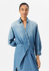 Denim omslagskjole med krage, lange ermer og knappedetaljer. Lys blå farge med myk tekstur og rynket midje.