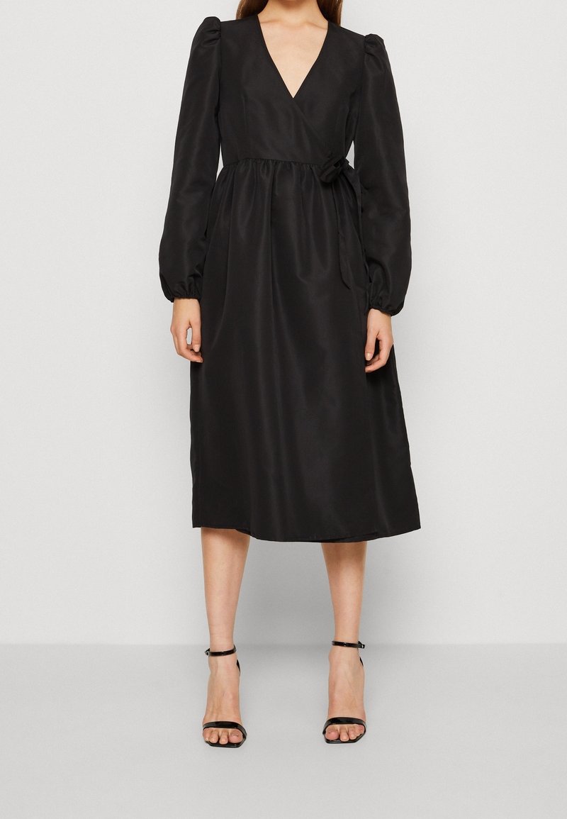 Femme portant une robe portefeuille midi noire avec de longues manches bouffantes et des sandales à talons hauts noires avec bride à la cheville, debout devant un fond uni.