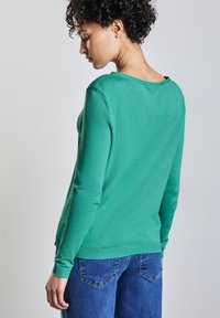 Frau mit kurzen, lockigen Haaren, die einen grünen Pullover mit langen Ärmeln und blaue Jeans trägt, seitlich vor einem einfarbigen Hintergrund.