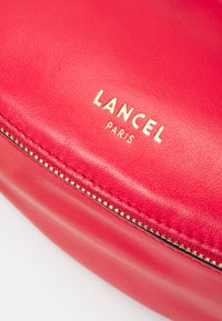 Sac en cuir rouge avec le logo doré "LANCEL PARIS" et un détail de fermeture éclair. Texture lisse avec des coutures visibles le long du bord de la fermeture éclair.