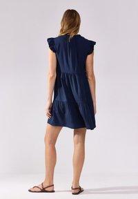 Abito blu navy con maniche corte a volant, design a strati e tessuto morbido. La vista posteriore mostra una lunghezza sopra il ginocchio e dettagli minimi.