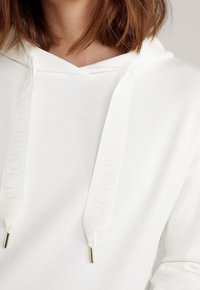 Gros plan d'une personne portant un sweat à capuche blanc avec des cordons larges et texturés comportant un texte embossé subtil.