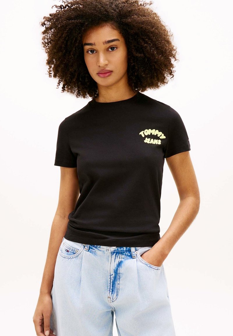 Jeune femme aux cheveux bouclés portant un t-shirt noir "Tommy Jeans" et un jean en denim bleu clair, debout avec une main dans la poche.