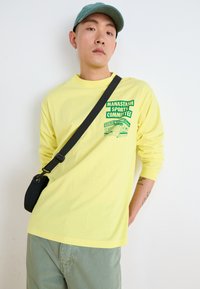 Camisa amarilla de manga larga con un estampado gráfico verde en el lado izquierdo del pecho. Combinada con pantalones verdes claros y una bolsa de hombro negra.