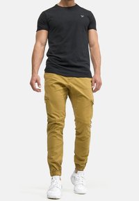 Schwarzes T-Shirt mit kurzen Ärmeln, taillierter Schnitt, kombiniert mit senfgelben Cargo-Hosen mit Reißverschlusstaschen. Weiße Sneaker runden den Look ab.